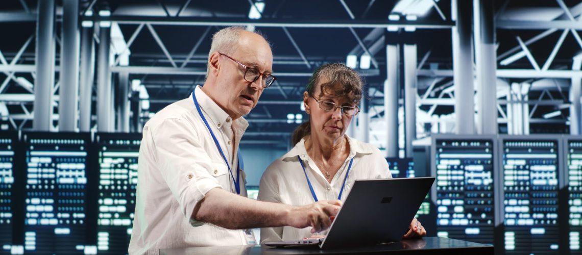 Senescent colleagues looking around high tech data center, using laptop to crosscheck disaster recovery plan and assess server machines in need of replacement, preventing glitches